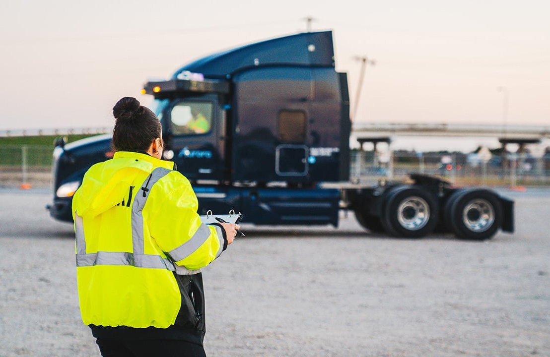 A woman in a yellow jacket facing way from the camera standing in front of a semi-truck. 