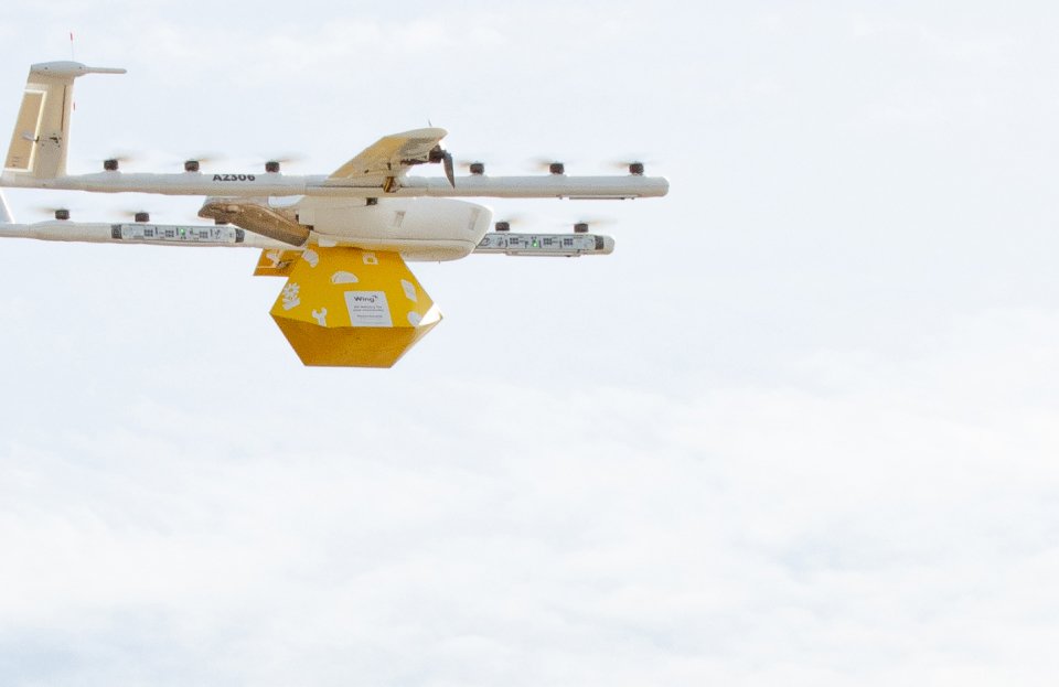 A Wing Aviation drone carrying a yellow package through the sky.