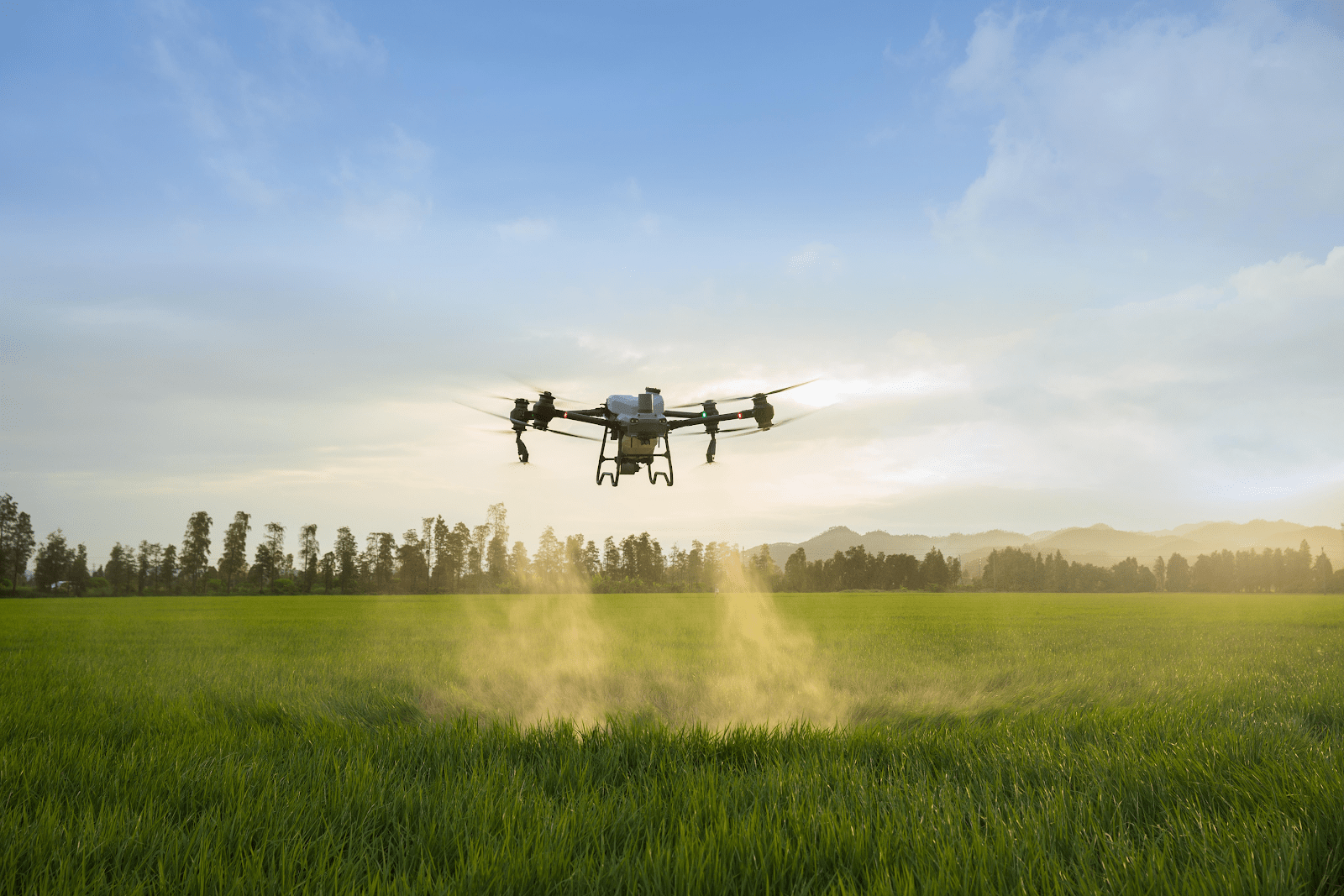 A drone hovers over a green field, dispersing a mist, likely for agricultural capabilities, with a background of bushes and far-off mountains throughout daylight.