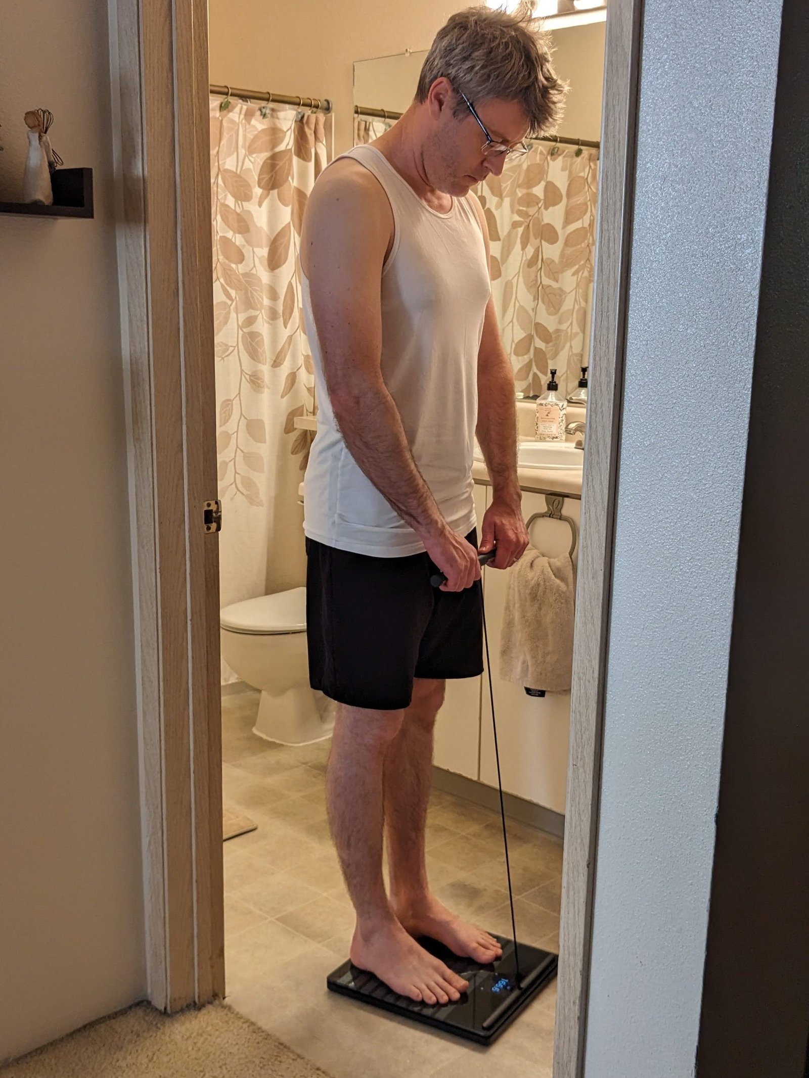 A individual in a white tank top and sad shorts stands on a scale in a lavatory, attempting down on the scale's prove.