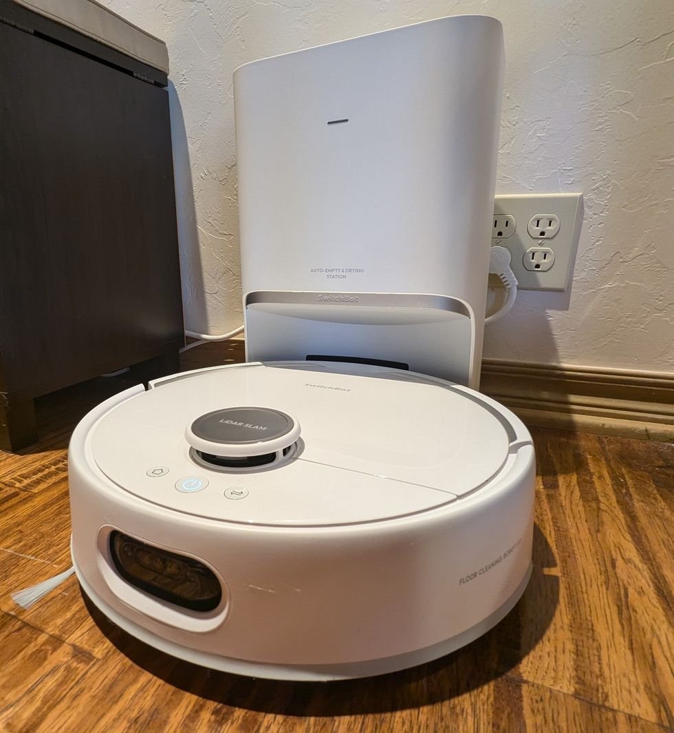 A large white rectangular dock sits plugged in against a wall with a round white robot vacuum docked to it.