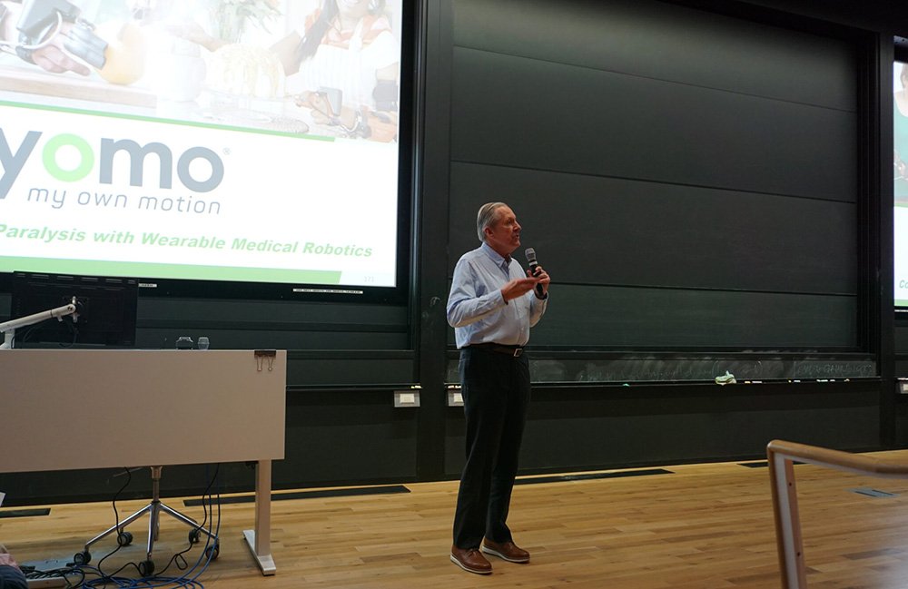 Paul Gudonis, CEO of Myomo, standing on a stage with a projector behind him, giving a talk at a recent MassRobotics event about robots for aging.