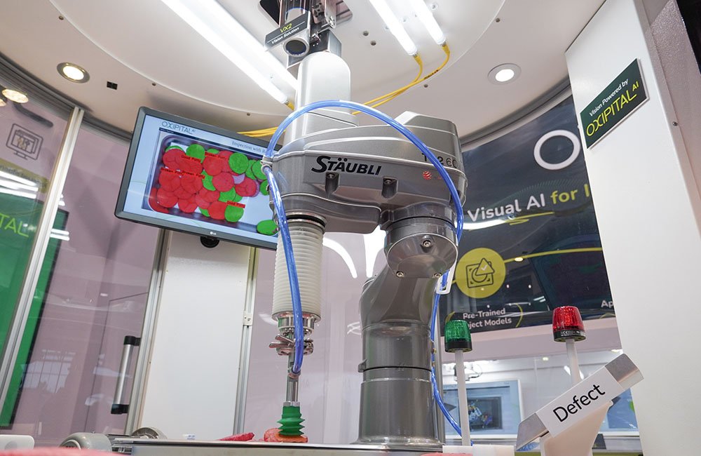 A grey Stäubli SCARA robot with a green suction cup end effector picking a hamburger patty using Oxipital AI's software. 