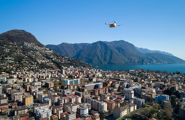 A white Matternet drone flying above a dense city with mountains in the background. 