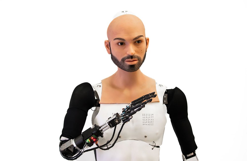 A Realbotix humanoid robot, from chest up. It's head and neck have a hyperrealistic covering, while its chest and arms are normal robot parts. 