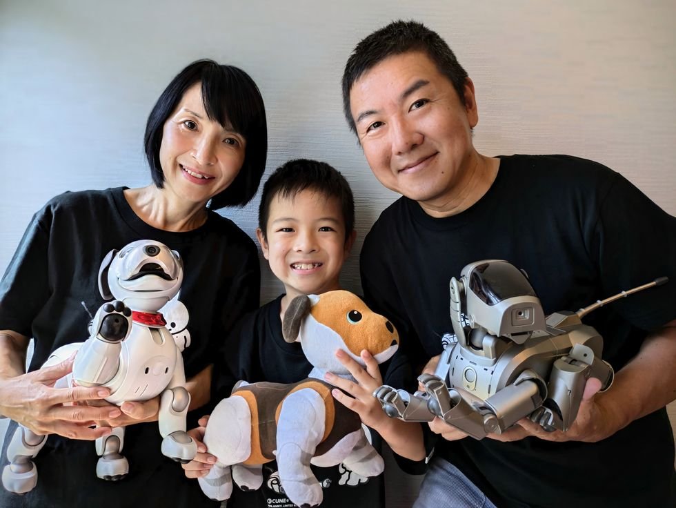 Smiling middle aged Japanese parents and their young son are all holding robot dogs, the mother holds a modern robot dog, the father holds an older robot dog, and the son holds a stuffed robot dog.