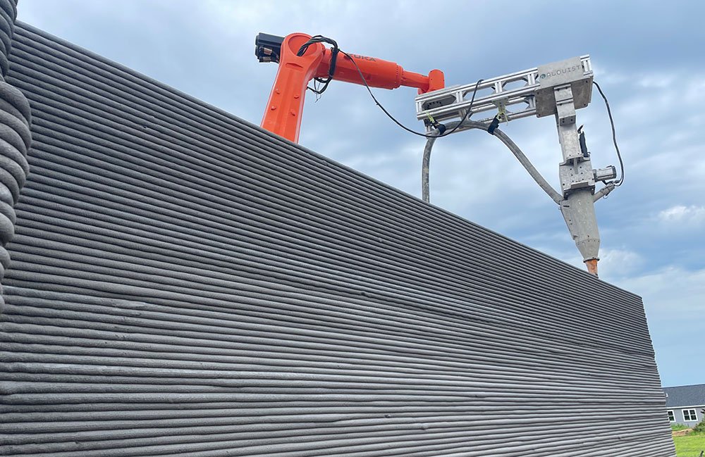 An orange KUKA robot with a custom attachment 3D printing a concrete house. 